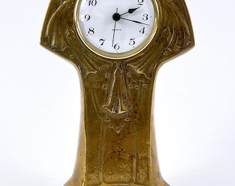 Op de afbeelding: Een messingkleurige quartz klok met een witte wijzerplaat en zwarte wijzers en cijfers. De klok heeft een Art Nouveau-ontwerp met bloemen en vloeiende lijnen. De klok is ongeveer 15 cm hoog.