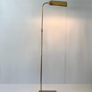 Puede incluir: Una l&aacute;mpara de pie de lat&oacute;n con una base rectangular y un poste vertical y delgado. La l&aacute;mpara tiene un brazo curvo que sostiene una l&aacute;mpara rectangular. La l&aacute;mpara est&aacute; enchufada a una toma de corriente con un interruptor blanco.
