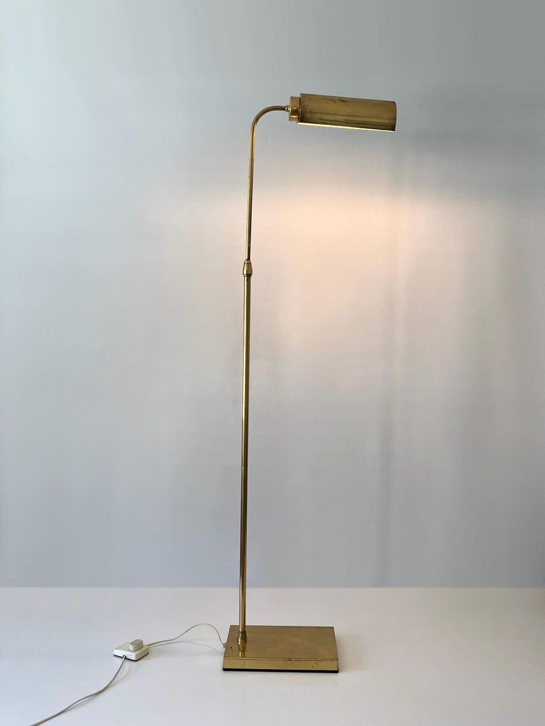Puede incluir: Una l&aacute;mpara de pie de lat&oacute;n con una base rectangular y un poste vertical y delgado. La l&aacute;mpara tiene un brazo curvo que sostiene una l&aacute;mpara rectangular. La l&aacute;mpara est&aacute; enchufada a una toma de corriente con un interruptor blanco.