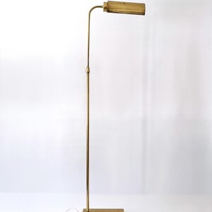 Puede incluir: Una l&aacute;mpara de pie vintage de lat&oacute;n con una base rectangular y un tallo largo y delgado. La l&aacute;mpara tiene una pantalla rectangular horizontal. La l&aacute;mpara est&aacute; enchufada a una toma de corriente. El dise&ntilde;o es minimalista y elegante.