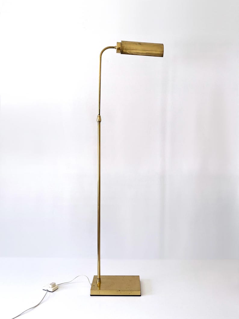 Puede incluir: Una l&aacute;mpara de pie vintage de lat&oacute;n con una base rectangular y un tallo largo y delgado. La l&aacute;mpara tiene una pantalla rectangular horizontal. La l&aacute;mpara est&aacute; enchufada a una toma de corriente. El dise&ntilde;o es minimalista y elegante.