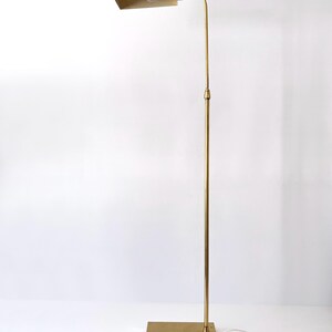 Puede incluir: L&aacute;mpara de pie de lat&oacute;n vintage con una pantalla rectangular y un brazo curvo. La l&aacute;mpara tiene una base cuadrada y un poste alto y delgado. La l&aacute;mpara est&aacute; enchufada a una toma de corriente blanca. Dise&ntilde;o moderno de mediados de siglo.