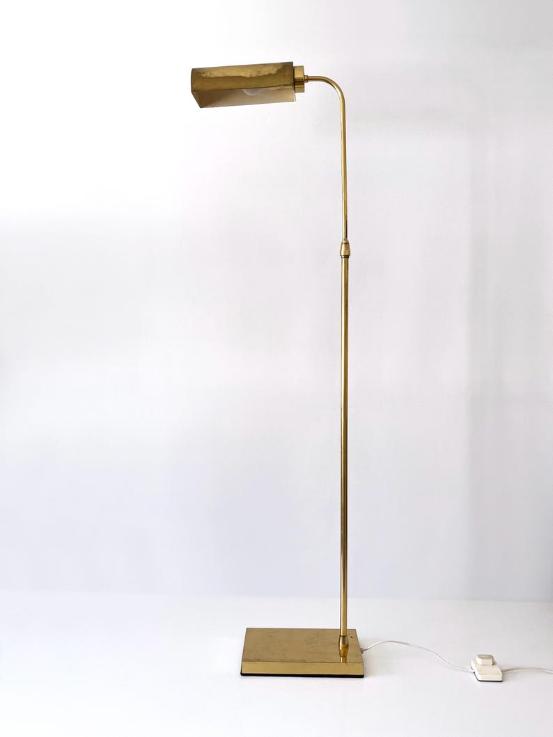 Puede incluir: L&aacute;mpara de pie de lat&oacute;n vintage con una pantalla rectangular y un brazo curvo. La l&aacute;mpara tiene una base cuadrada y un poste alto y delgado. La l&aacute;mpara est&aacute; enchufada a una toma de corriente blanca. Dise&ntilde;o moderno de mediados de siglo.