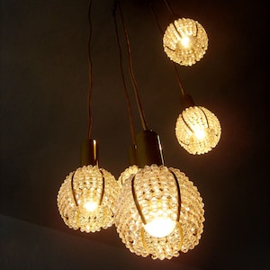 Op de afbeelding: Een gouden kroonluchter met vijf heldere kristallen bolvormige lampen. De kroonluchter hangt aan het plafond en de lampen zijn aan.