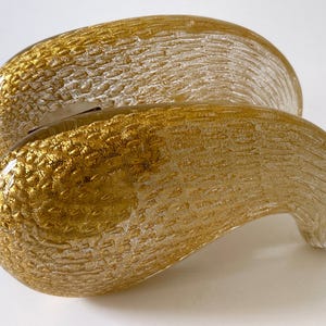 May include: An abstract glass sculpture in gold and clear tones. The piece has a textured surface and a curved, organic shape. Likely a decorative item for a home or office. The sculpture's dimensions are not visible.