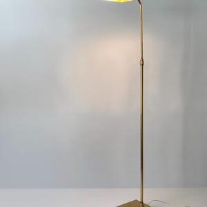Puede incluir: Una l&aacute;mpara de pie de lat&oacute;n con una base rectangular y una pantalla curva y rectangular. La l&aacute;mpara tiene un poste delgado y ajustable y un cable con un interruptor. La luz est&aacute; encendida, iluminando el interior de la pantalla.