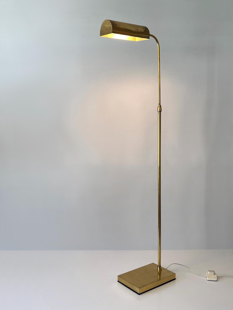Puede incluir: Una l&aacute;mpara de pie de lat&oacute;n con una base rectangular y una pantalla curva y rectangular. La l&aacute;mpara tiene un poste delgado y ajustable y un cable con un interruptor. La luz est&aacute; encendida, iluminando el interior de la pantalla.