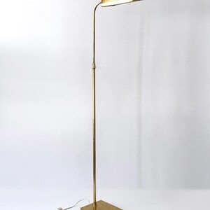 Puede incluir: Una l&aacute;mpara de pie de color lat&oacute;n con una base rectangular y un poste vertical y delgado. La l&aacute;mpara presenta una pantalla curva y rectangular que alberga una fuente de luz. La l&aacute;mpara est&aacute; enchufada a una toma de corriente mediante un cable blanco.