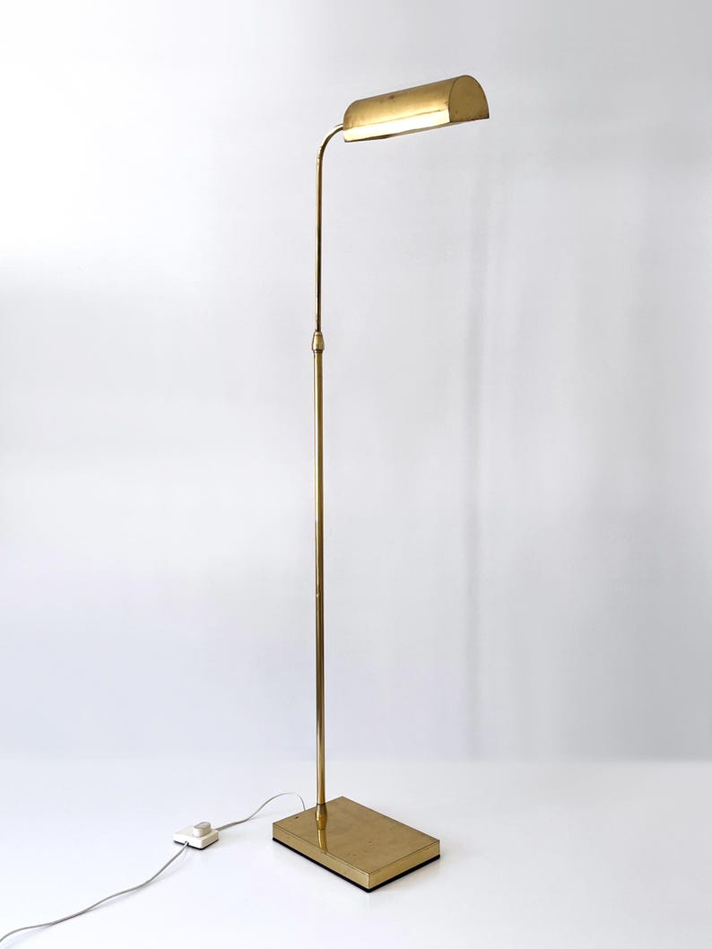 Puede incluir: Una l&aacute;mpara de pie de color lat&oacute;n con una base rectangular y un poste vertical y delgado. La l&aacute;mpara presenta una pantalla curva y rectangular que alberga una fuente de luz. La l&aacute;mpara est&aacute; enchufada a una toma de corriente mediante un cable blanco.