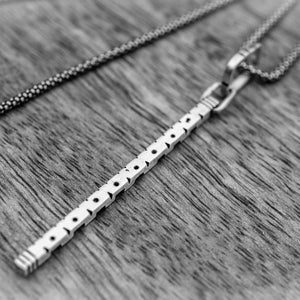 May include: A silver necklace with a rectangular pendant featuring a series of small, evenly spaced holes. The pendant is connected to a delicate chain. Another chain is visible in the background. The jewelry rests on a wooden surface.