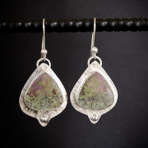 May include: A pair of silver earrings with a teardrop shape. Each earring features a textured silver frame surrounding a stone with green and purple hues. The earrings hang from silver hooks and have a small silver bead at the bottom.
