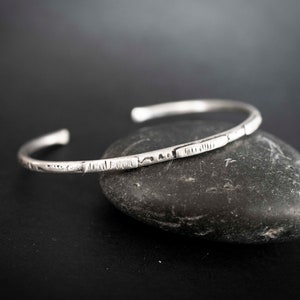 May include: A silver bangle bracelet with a hammered texture. The bracelet is open and has a simple design.