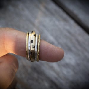 May include: A stack of five rings worn on a finger, featuring a mix of gold and silver-toned metal bands. The central band displays a repeating geometric pattern of small squares and triangles, while the surrounding bands have smooth or textured finishes.
