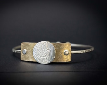 May include: A silver and gold bracelet with a textured, circular design in the center. The bracelet is made of metal and has a simple, elegant design.