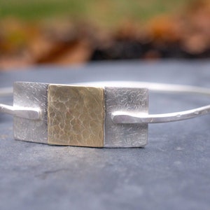 May include: A silver bangle bracelet with a rectangular gold-toned center piece. The center piece has a textured surface, flanked by two silver rectangles. The bracelet is displayed on a dark gray surface.