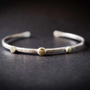 May include: A silver cuff bracelet with three small, gold-colored circular accents. The bracelet has a hammered texture and is open at the ends. The jewelry is displayed on a dark surface.