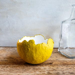 May include: A yellow paper mache bowl with a white interior and a rough, textured surface. The bowl is sitting on a wooden surface.