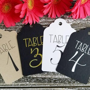 May include: Four decorative table number tags with scalloped edges. The tags are in various colours: tan, black, and white. Each tag has the word "TABLE" and a number written on it. Bright pink flowers are in the background.