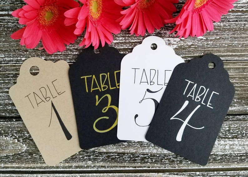 May include: Four decorative table number tags with scalloped edges. The tags are in various colours: tan, black, and white. Each tag has the word "TABLE" and a number written on it. Bright pink flowers are in the background.