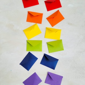 May include: A collection of colorful envelopes arranged in a rainbow spectrum. The envelopes are in pairs of red, orange, yellow, green, blue, and purple. The envelopes are on a light-colored surface.