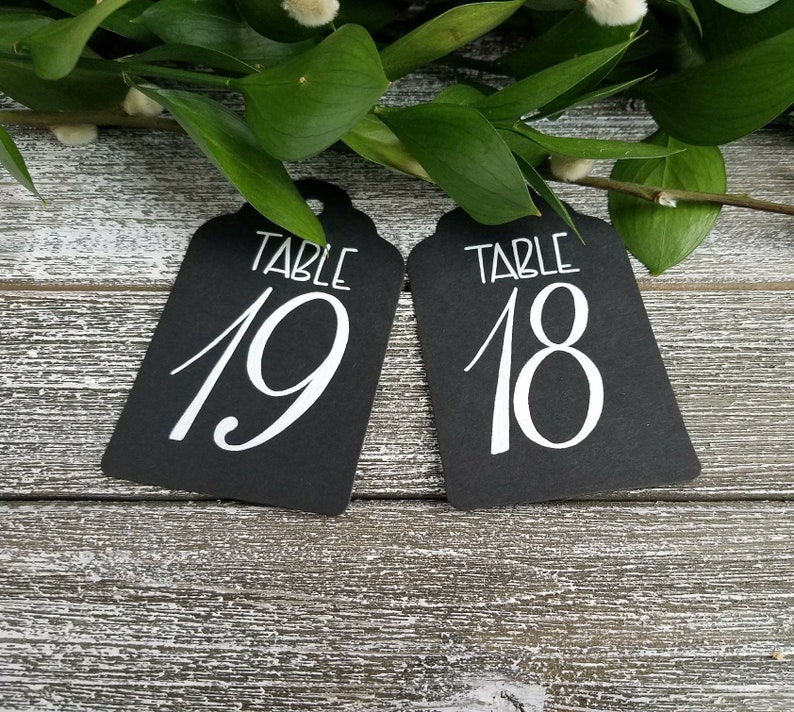 May include: Two black table number tags with white calligraphy. The tags are labelled "TABLE 19" and "TABLE 18". Greenery and a wooden surface provide a rustic backdrop. Ideal for weddings or events.