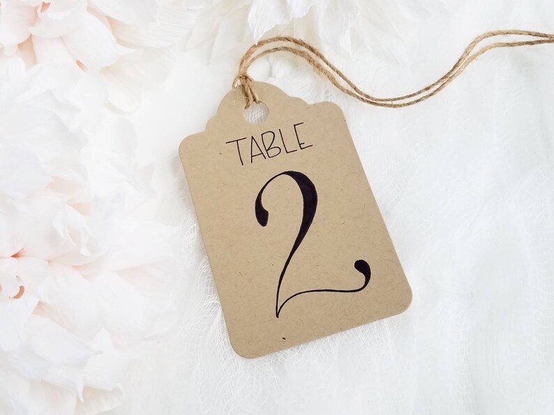 May include: A brown paper table number tag with the word "TABLE" and the number "2" in black script. The tag is attached to a twine string. The background is white with soft, white floral accents.