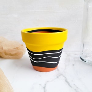 May include: A small, yellow ceramic flower pot with a black and white wavy design. The pot has a terracotta base and a black rim. The pot is empty and ready for planting.