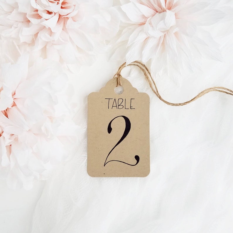 May include: A brown paper table number tag with the word "TABLE" and the number "2" in black script. The tag is suspended by a natural twine string. Soft pink flowers and white fabric create a delicate background.