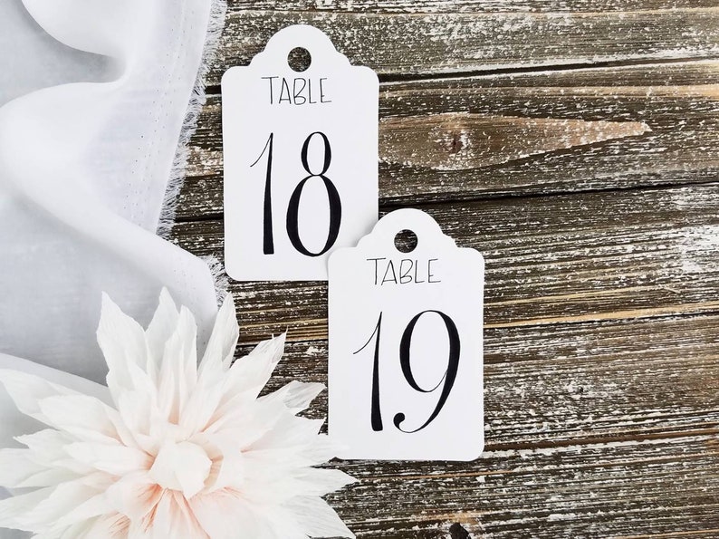 May include: Two white table number cards with a scalloped top, displaying the numbers 18 and 19 in black script. The word "TABLE" is above each number. A white flower and fabric are in the foreground.