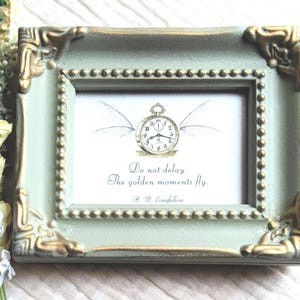 Puede incluir: Un pequeño marco de fotos rectangular en un tono verde suave con detalles dorados. El marco contiene una impresión con un reloj de bolsillo con alas y la cita "Do not delay. The golden moments fly." de H.W. Longfellow.