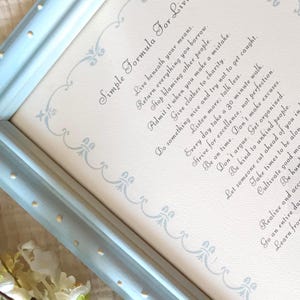 May include: A framed print with a light blue wooden frame. The print features a handwritten script with the title "Simple Formula For Living" and a list of life principles. The frame has small, decorative dots.