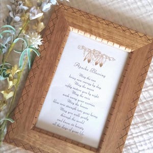 May include: A rectangular wooden frame with a cross-hatch pattern along the edges, containing a white card printed with the text Apache Blessing and a poem about the sun, moon, rain, and breeze. The frame is light brown with a visible wood grain.