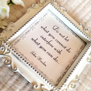 Op de afbeelding: Een vierkante, sierlijke wit-zilveren lijst met een crèmekleurige kaart waarop in zwarte letters staat: Do not let what you cannot do interfere with what you can do.. John Wooden. De lijst is voorzien van gedetailleerde krul- en kraalmotieven.