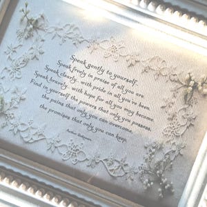 May include: A framed piece of art with a white, decorative frame. The artwork displays a quote in elegant script, surrounded by delicate floral and lace details. The quote starts with, "Speak gently to yourself..." and is by "Author Unknown."
