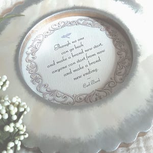 May include: A decorative, scalloped-edge plate with a central circular design. The design features an ornate border and a quote by Carl Bard: "Though no one can go back and make a brand new start..." The plate is a soft, muted color.