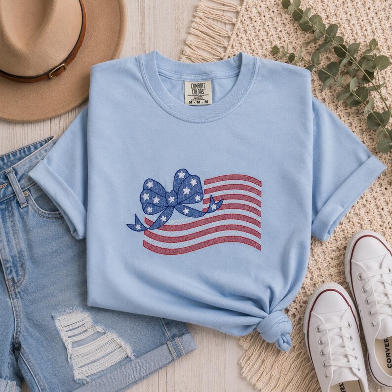 May include: A light blue t-shirt featuring an embroidered American flag design. The flag has red and white stripes with a blue bow adorned with white stars. The shirt is a Comfort Colors brand and is displayed with denim shorts and white sneakers.