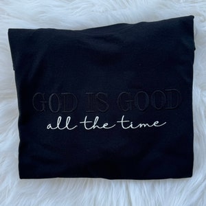 K&ouml;nnte beinhalten: Schwarzes T-Shirt mit dem gestickten Text "GOD IS GOOD all the time" in Wei&szlig;.