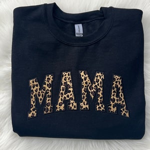 Puede incluir: Sudadera negra con la palabra "MAMA" en letras grandes, en negrita y con estampado de leopardo.