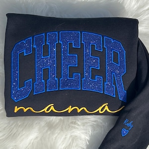 Könnte beinhalten: Schwarzer Sweatshirt mit blauem Glitzer-Text "CHEER mama". Der Sweatshirt hat einen goldenen Glitzer-Text "mama" und ein kleines blaues Herz mit dem Text "Ruby" auf dem Ärmel.
