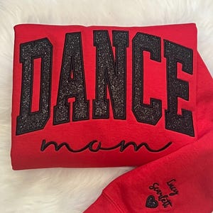 May include: Red sweatshirt with black glitter lettering that says "Dance Mom". The sweatshirt has a small heart with the name "Lucy Scarlett" on the sleeve.