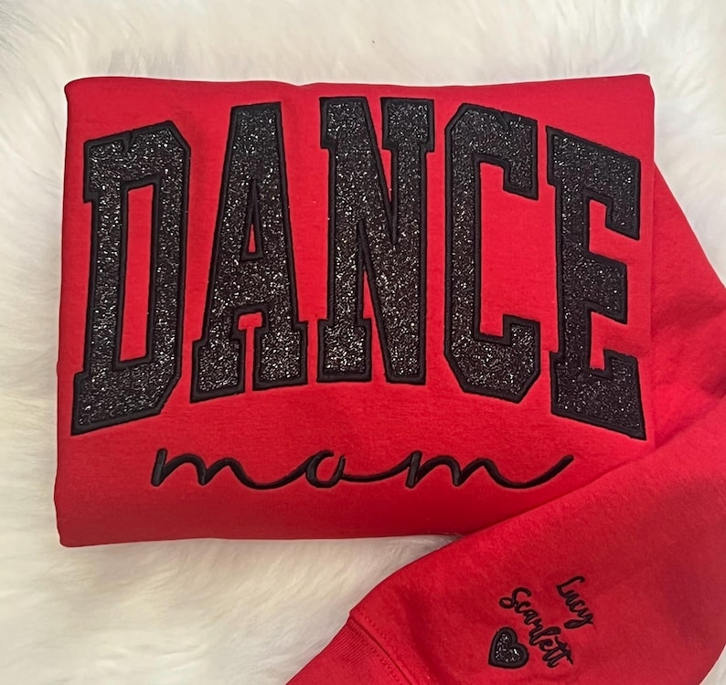 May include: Red sweatshirt with black glitter lettering that says "Dance Mom". The sweatshirt has a small heart with the name "Lucy Scarlett" on the sleeve.