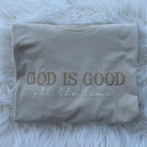 K&ouml;nnte beinhalten: Ein beiges T-Shirt mit dem Text "GOD IS GOOD all the time", der in hellbrauner Farbe gestickt ist.