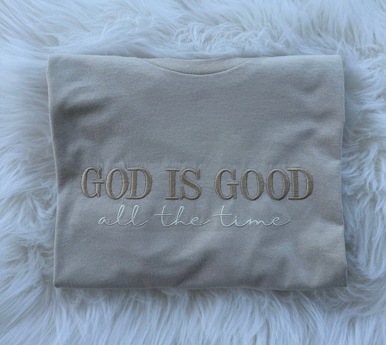K&ouml;nnte beinhalten: Ein beiges T-Shirt mit dem Text "GOD IS GOOD all the time", der in hellbrauner Farbe gestickt ist.