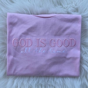 K&ouml;nnte beinhalten: Rosa T-Shirt mit dem gestickten Text "GOD IS GOOD all the time" in Rosa und Wei&szlig;.