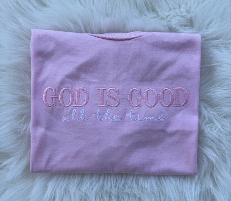 K&ouml;nnte beinhalten: Rosa T-Shirt mit dem gestickten Text "GOD IS GOOD all the time" in Rosa und Wei&szlig;.
