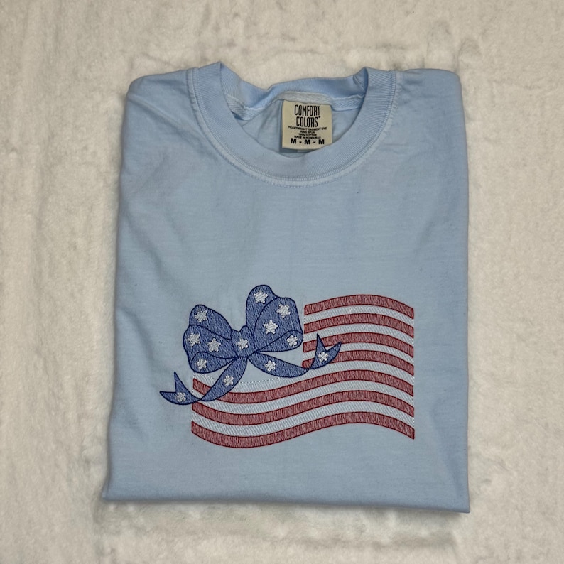 May include: Light blue t-shirt with an embroidered American flag design. The flag features red and white stripes and a blue bow with white stars. The shirt is folded and has a Comfort Colors tag.