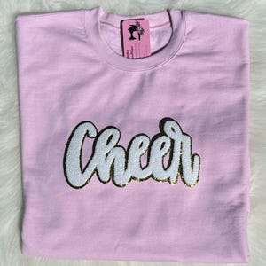 Puede incluir: Sudadera rosa con la palabra "Cheer" en blanco con letras doradas con purpurina.