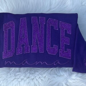 May include: Purple sweatshirt with the text "DANCE mama" in sparkly purple letters. The sweatshirt has a small heart with the name "Emma" embroidered on the bottom right corner.