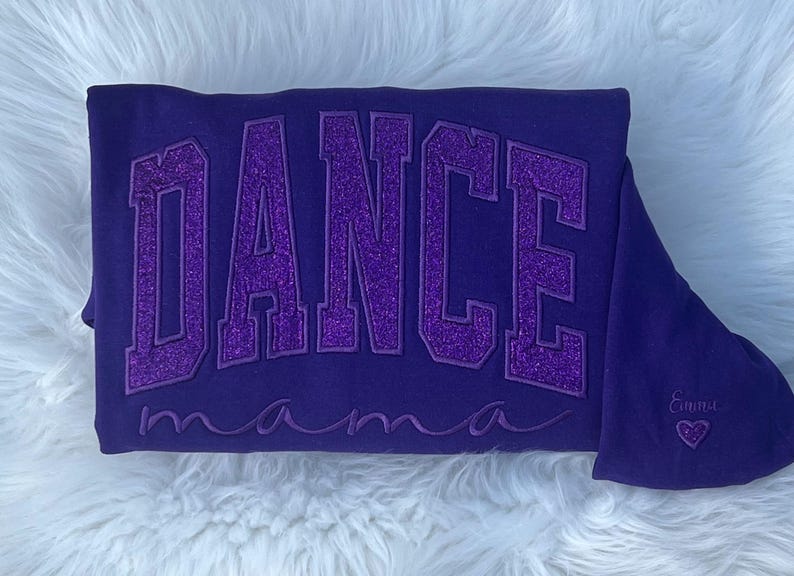 May include: Purple sweatshirt with the text "DANCE mama" in sparkly purple letters. The sweatshirt has a small heart with the name "Emma" embroidered on the bottom right corner.