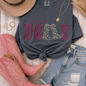 Könnte beinhalten: Ein anthrazitfarbenes Kurzarm-T-Shirt mit dem Text "raised on DOLLY" in Pink und Gelb sowie Cowboystiefel-Grafiken. Dazu kombiniert sind pinke Cowboystiefel, hellblaue Jeansshorts, ein beigefarbener Hut und eine goldene Anhängerkette.